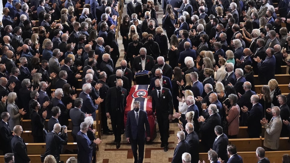 People in a cathedral applaud the passage of a coffin covered with a sheet in the colors of the Montreal Canadiens.