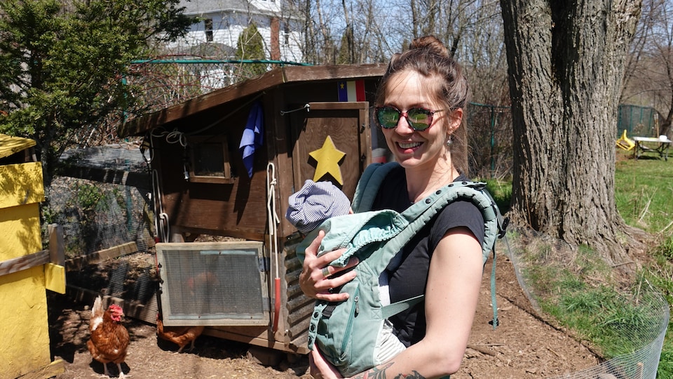 A young woman with a baby in her arms stands in front of a small chicken coop in a house yard. 
