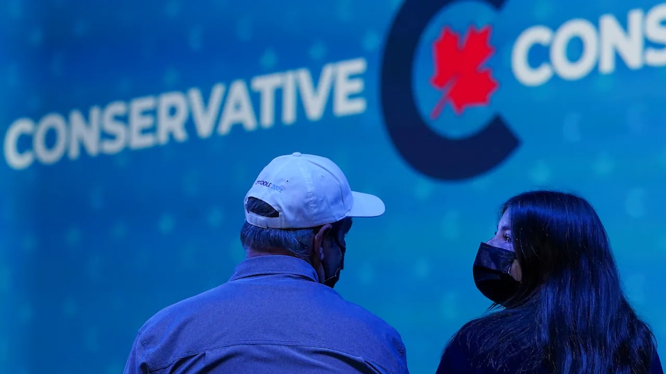 A masked man and woman discuss in front of a giant screen in the colors of the Conservative Party of Canada.