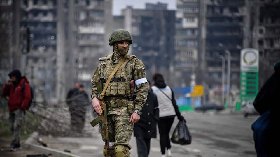 A Russian soldier in the middle of a street.  In the background, people are walking down a street.  Charred apartment buildings are visible. 