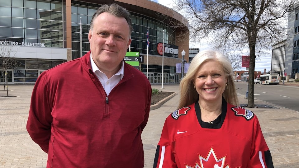 Mike Savage wears a red jersey and Dawn Arnold wears a Team Canada hockey jersey.  They both pose outside in front of an arena.