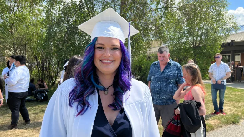 Calysta Stoker smiles with purple hair and wearing a white grad robe for her graduation.