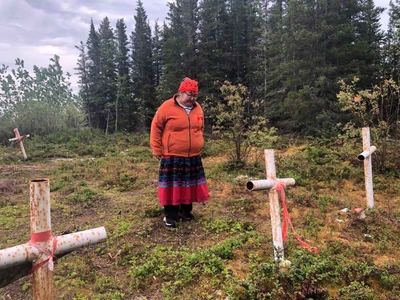 A woman wearing an orange sweatshirt and a multi-coloured ribbon skirt stands near a grave on rough terrain.