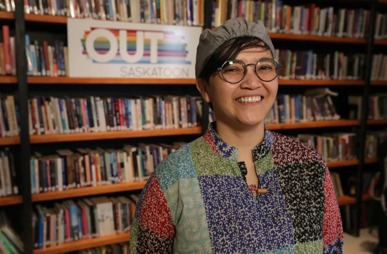 A person smiles for the camera in front of a bookcase with a sign reading 'OUTSaskatoon'. 