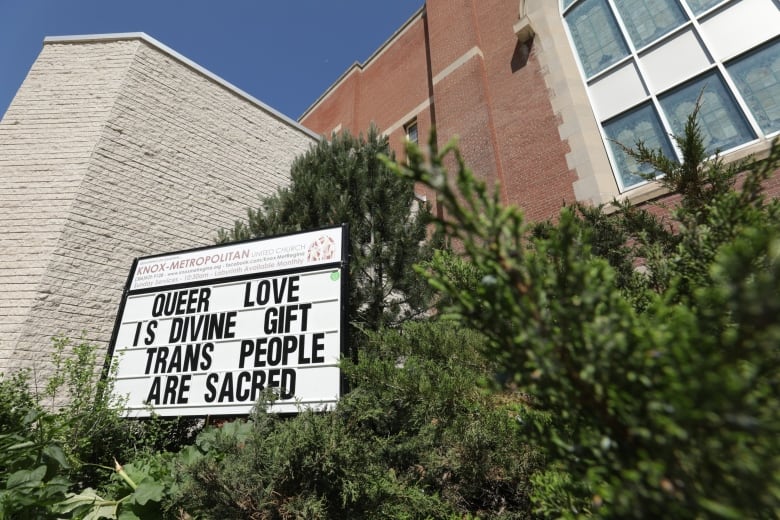 A sign that says 'Queer love is divine gift; trans people are sacred' sits amongst evergreen bushes outside of a church.