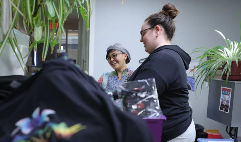 Two people organize Pride merchandise, including black shirts printed with rainbow lilies, in the OUTSaskatoon office.