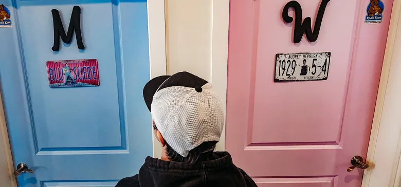 A person stands in front of two washroom doors: the one on the left, which they are looking at, is labelled with an 'M' and the one on the right is pink, labelled with a 'W'.