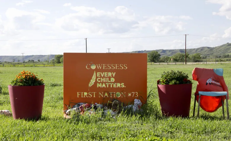 An orange sign with the words "Cowessess, Every Child Matters. First Nation #73" sit in a field. Stuffed animals rest at the base of the sign, which is flanked by two potted plants and a chair with an orange shirt on it. 