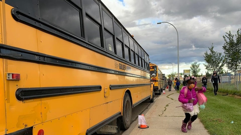A student has just got off a school bus.  She wears a yellow mask and walks towards the entrance of Father Leo Green Elementary School in Edmonton.  A group of other people are behind her.