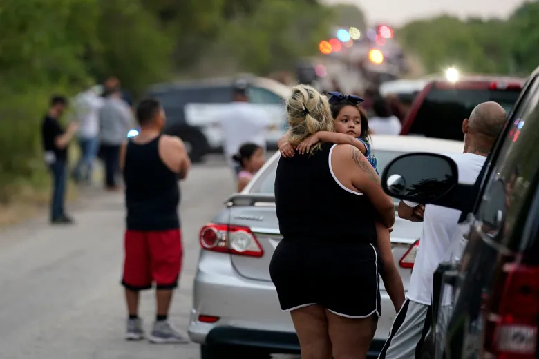 Two individuals, one holding a child, are seen from behind, looking at the truck and emergency personnel. 