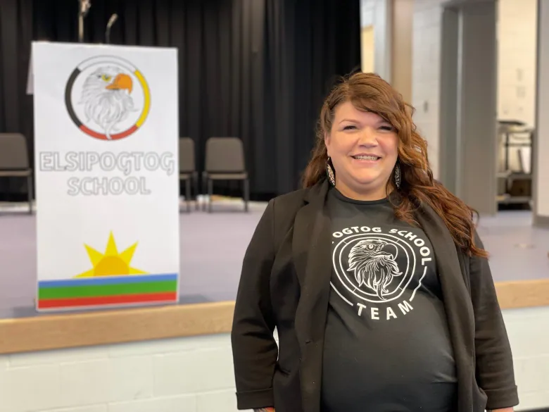 Melissa Googoo Dedam, princpal of Elsipogtog First Nation Community School, stands smiling in the school auditorium in front of a stage and podium that says Elsipogtog School.
