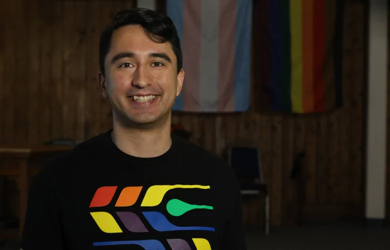 A man smiles inside a church. He is standing in front of two Pride flags and wearing a Pride T-shirt with a rainbow feather design.
