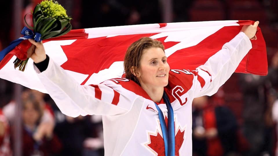 Hayley Wickenheiser with her Olympic gold medal and the Canadian flag above her head on the ice in Vancouver